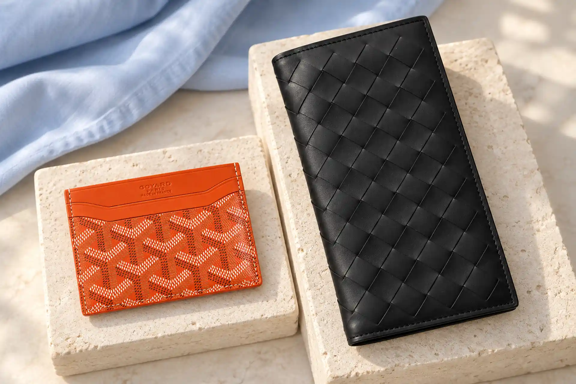 Оранжевый картхолдер Goyard и чёрный плетёный купюрник Bottega Veneta на светлых подставках — купить брендовую одежду и аксессуары.